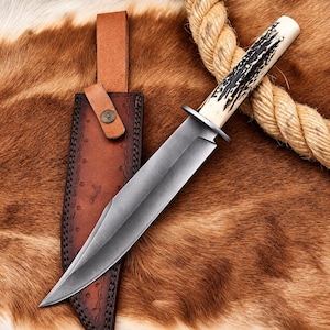 May include: A fixed-blade knife with a dark grey steel blade and a textured, cream-colored handle featuring dark brown accents. The knife includes a brown leather sheath with decorative stitching and a metal snap button closure for secure storage.