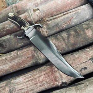 May include: A large, stainless steel Bowie knife with a polished blade and a handle made of antler material. The knife features a curved blade and a guard. The knife is displayed on a bamboo surface.