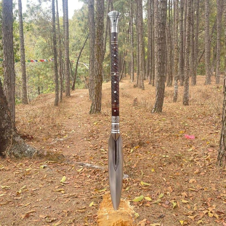May include: A long, ornate spear with a silver blade and a dark wooden shaft. The shaft features a decorative pattern of silver and white accents. The spear is standing upright, with the blade resting on a wooden surface, set against a backdrop of a forest.