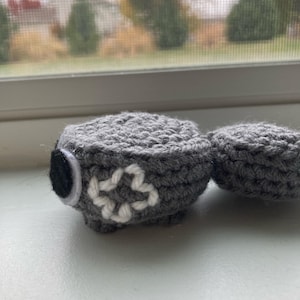 Op de afbeelding: Handgemaakt grijs gehaakt item met een wit bloemmotief en een zwart cirkelvormig accent. Het item ligt op een wit oppervlak bij een raam.
