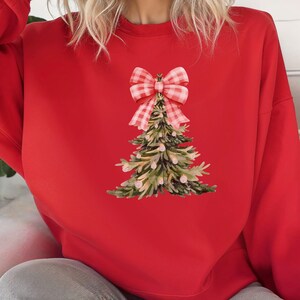 Peut inclure: Un sweat-shirt rouge avec un motif d'arbre de Noël à l'aquarelle et un nœud vichy rose et blanc. L'arbre a des aiguilles vertes et marron avec des accents roses. Le sweat-shirt est de couleur rouge uni.
