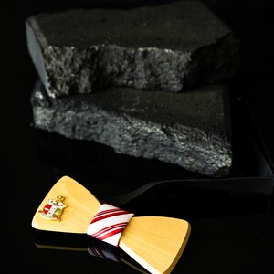 Pode incluir: Uma gravata borboleta de madeira com acabamento castanho claro, com um centro listrado em vermelho e branco e um emblema dourado. A gravata borboleta está sobre um fundo preto com dois blocos de pedra escuros e texturizados.
