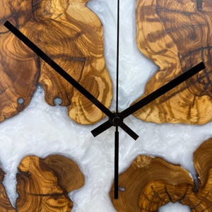 K&ouml;nnte beinhalten: Eine Holz-Wanduhr mit einzigartigem Design. Das Zifferblatt zeigt Abschnitte aus hellbraunem Holz mit komplizierten Maserungsmustern, durchsetzt mit wei&szlig;em, flie&szlig;endem Harz. Schwarze Zeiger zeigen die Uhrzeit an.