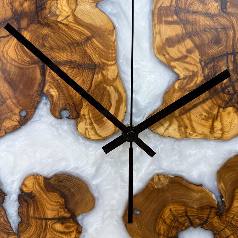 K&ouml;nnte beinhalten: Eine Holz-Wanduhr mit einzigartigem Design. Das Zifferblatt zeigt Abschnitte aus hellbraunem Holz mit komplizierten Maserungsmustern, durchsetzt mit wei&szlig;em, flie&szlig;endem Harz. Schwarze Zeiger zeigen die Uhrzeit an.