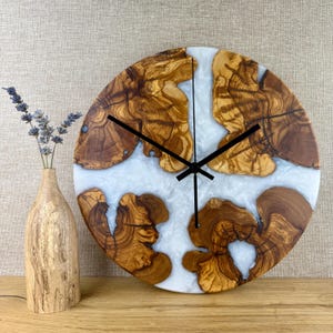 K&ouml;nnte beinhalten: Eine runde Wanduhr mit einzigartigem Design. Das Zifferblatt zeigt Abschnitte mit nat&uuml;rlicher Holzmaserung und wei&szlig;em, marmoriertem Harz. Schwarze Zeiger zeigen die Uhrzeit an. Links steht eine Holzvase mit Lavendel.