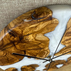 K&ouml;nnte beinhalten: Eine handgefertigte Holzuhr mit einzigartigem Design. Das Zifferblatt zeigt eine Kombination aus nat&uuml;rlicher Holzmaserung und wei&szlig;em Harz. Die Zeiger sind schwarz, und die Uhr hat einen Durchmesser von etwa 30 cm.