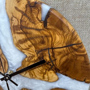 K&ouml;nnte beinhalten: Eine Holz-Uhr mit einem nat&uuml;rlichen Holzmaserungsmuster und einem wei&szlig;en Harzeinlagen. Die Uhr hat schwarze Zeiger und eine goldfarbene Mitte. Das Holz hat einen warmen, braunen Ton, und das Harz sorgt f&uuml;r einen modernen Kontrast.