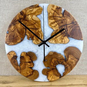 K&ouml;nnte beinhalten: Runde Wanduhr aus Olivenholz und hellem Epoxidharz. Die Uhr hat ein nat&uuml;rliches Holzmaserungsmuster und schwarze Zeiger. Ein dekorativer Artikel f&uuml;r das Zuhause, mit einem Durchmesser von etwa 30 cm.