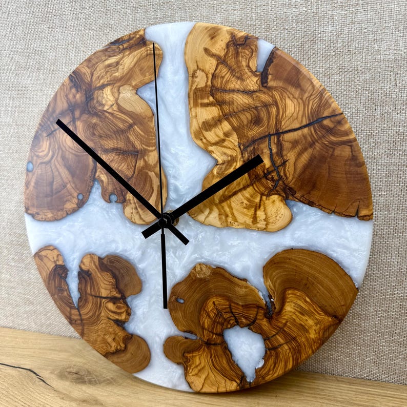 K&ouml;nnte beinhalten: Runde Wanduhr aus Holz und Kunstharz. Das Zifferblatt zeigt nat&uuml;rliche Holzmaserungen in Braun, unterbrochen von wei&szlig;em Kunstharz. Schwarze Zeiger zeigen die Uhrzeit an. Der Durchmesser betr&auml;gt ca. 25 cm.