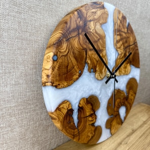 K&ouml;nnte beinhalten: Runde Wanduhr aus Olivenholz und Kunstharz. Das Zifferblatt hat hellfarbenes Kunstharz, das die Zwischenr&auml;ume zwischen den Holzabschnitten ausf&uuml;llt. Schwarze Zeiger und ein goldfarbener Mittelpunkt vervollst&auml;ndigen das Design. Die Uhr ist an einer neutralfarbenen Wand befestigt.