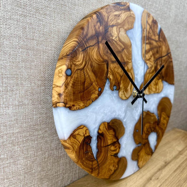 K&ouml;nnte beinhalten: Runde Wanduhr aus Olivenholz und Kunstharz. Das Zifferblatt hat hellfarbenes Kunstharz, das die Zwischenr&auml;ume zwischen den Holzabschnitten ausf&uuml;llt. Schwarze Zeiger und ein goldfarbener Mittelpunkt vervollst&auml;ndigen das Design. Die Uhr ist an einer neutralfarbenen Wand befestigt.