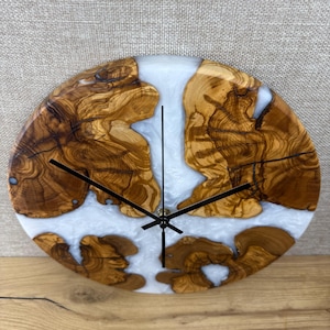 K&ouml;nnte beinhalten: Eine runde Wanduhr mit einem einzigartigen Design. Das Zifferblatt zeigt Abschnitte aus Naturholz mit einem hellfarbigen Harz, das die L&uuml;cken ausf&uuml;llt. Schwarze Zeiger zeigen die Uhrzeit an. Das Holz hat eine satte, braune Farbe.