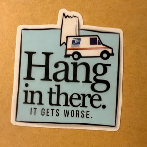May include: A sticker with a light blue background and the words "Hang in there. IT GETS WORSE." in black. A white United States Postal Service truck is at the top. The sticker has a white border and is on a brown surface.