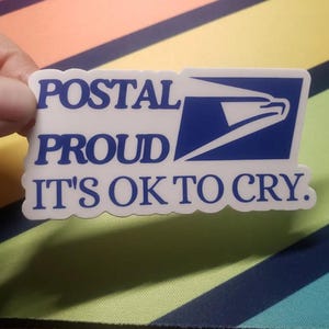 May include: A white sticker with blue text and a blue postal service logo. The text reads "POSTAL PROUD IT'S OK TO CRY."