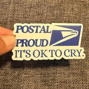 May include: A white sticker with blue text and a blue postal service logo. The text reads "POSTAL PROUD IT'S OK TO CRY."