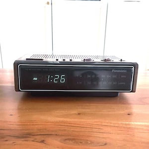 May include: A vintage Panasonic digital clock radio with a dark wood-grain finish. The clock displays 1:26 AM in green digital numbers. The radio has FM and AM radio frequency indicators and a speaker grill on top.