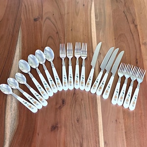 May include: A set of vintage cutlery with white handles and a pineapple motif. The collection includes spoons, forks, and knives, displayed on a wooden surface. The cutlery has a silver finish and a decorative pattern.