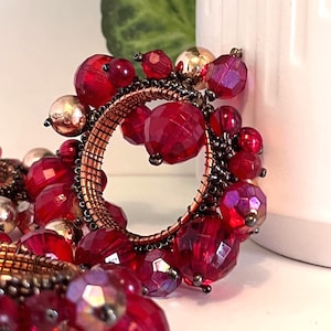 May include: A decorative circular ornament featuring faceted red beads and gold-colored beads. The beads are attached to a copper-colored wire frame. The ornament is placed near a white ceramic object and a green plant.