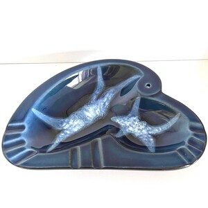 May include: A dark blue ceramic ashtray with a glossy finish, featuring a curved, asymmetrical shape and a central relief design of two textured, light blue starfish. The piece includes integrated notches along the rim for resting items.