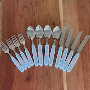 May include: A collection of vintage cutlery, comprising forks, spoons, and knives. The white handles feature a blue floral pattern with pink roses. The silverware is displayed on a wooden surface.
