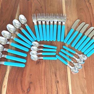 May include: A flatware set consisting of silver-toned metal forks, spoons, and knives, each featuring bright turquoise handles with a textured, ribbed grip pattern. The collection includes multiple pieces of each utensil type arranged in a fan-like display.