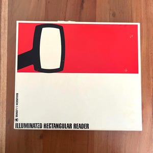 May include: A rectangular box with a red and white design. The top half is red with a black and white graphic. The bottom half is white with the text "ILLUMINATED RECTANGULAR READER" and the brand name "BAUSCH & LOMB".