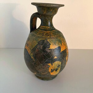 May include: An antique, olive green and orange ceramic vase with a handle. The vase features a black illustration of a person and geometric patterns. The vase is approximately 8 inches tall.