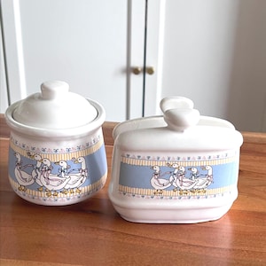 May include: Two white ceramic containers with lids. Each container features a blue band with a pattern of geese wearing pink bows. The containers are on a wooden surface.