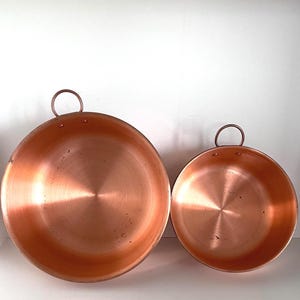 May include: Two copper pans with rounded sides and a circular handle. The pans are different sizes, with a shiny, reflective surface. The copper has a warm, reddish-brown hue, and the handles are attached with rivets.