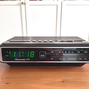 May include: Black and silver vintage Chronomatic-274 digital alarm clock. The time 11:18 is displayed in green. Features FM/AM radio, alarm, and battery sentinel. The clock sits on a wooden surface.