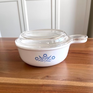 May include: White casserole dish with a clear glass lid and a handle. The dish features a blue floral design. The dish is sitting on a wooden surface.