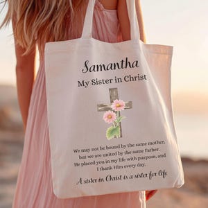 Op de afbeelding: Een crèmekleurige draagtas met de naam "Samantha" en de tekst "My Sister in Christ" in zwart script. Een kruis met roze bloemen is gecentreerd onder de tekst. Extra tekst is eronder gedrukt.