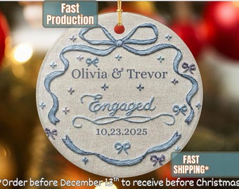 May include: A round, off-white Christmas ornament with blue embroidered details. The ornament features the names "Olivia & Trevor", the word "Engaged", and the date "10.23.2025". Decorative bows and ribbons adorn the edges.