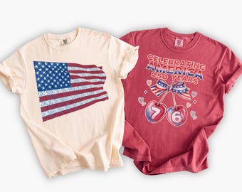 May include: Two t-shirts with patriotic designs. One is cream-colored with an American flag graphic and the number "76" repeated. The other is red with the text "Celebrating America 250 Years" and cherry graphics.
