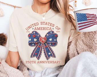 May include: Cream-colored t-shirt with a patriotic bow design. The bow is blue with red and white accents, featuring hearts and stars. Text includes "UNITED STATES OF AMERICA" and "250TH ANNIVERSARY". A mug with an American flag design is also visible.