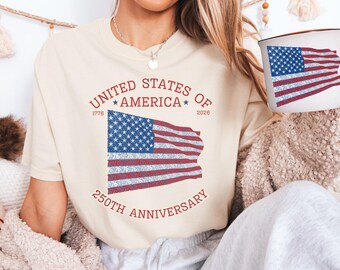 May include: Cream-colored t-shirt with a distressed American flag graphic. The design includes the text "UNITED STATES OF AMERICA" and "250TH ANNIVERSARY" with the years 1776 and 2026. A matching mug with the flag design is also visible.