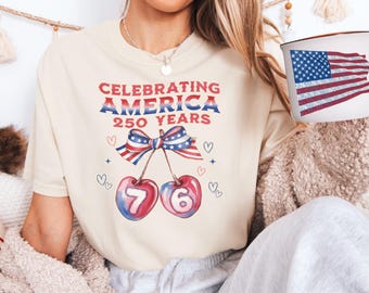 May include: Cream-colored t-shirt with the text "Celebrating America 250 Years" in red and blue. The design includes a bow with the American flag and cherries with the numbers 76. A mug with an American flag design is also visible.