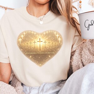Puede incluir: Camiseta color crema con un diseño en forma de corazón con cruces y el texto "Yesterday Today Forever". Se sostiene una taza blanca con la frase "God is good".