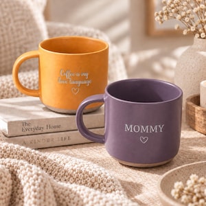 Op de afbeelding: Een gele keramische mok met de tekst Coffee is my love language en een harticoon, en een paarse keramische mok met de tekst MOMMY en een harticoon. Beide mokken hebben een gladde afwerking, een gebogen handvat en een lichtgekleurde onderkant.