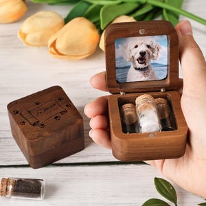 Puede incluir: Una caja conmemorativa de madera para mascotas con una foto de un perro en su interior. La caja está abierta, revelando pequeños frascos de vidrio con tapones de corcho. También se ve una caja separada con huellas de patas y texto.
