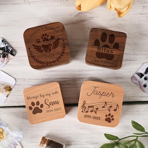 May include: Four wooden pet memorial boxes with engraved designs. Two are dark brown with paw prints, names, and dates. The other two are light brown, one with a paw print and the text "Always by my side", and the other with musical notes and a paw print.
