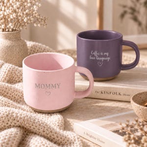 Könnte beinhalten: Ein hellrosa Keramikbecher mit dem Wort MOMMY und einem kleinen Herzsymbol auf der Vorderseite, gepaart mit einem dunkelvioletten Keramikbecher mit dem Text Coffee is my love language und einem kleinen Herzsymbol auf der Vorderseite.