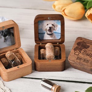 Op de afbeelding: Houten gedenkdozen voor huisdieren met fotolijsten en kleine glazen flesjes. Eén doos heeft een kattenfoto, een andere een hond, en de derde is gegraveerd met een honden silhouet, de naam "Daisy" en de data 2018-2025.
