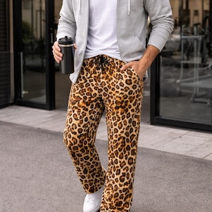 May include: A person wearing a gray zip-up hoodie, a white t-shirt, and leopard print pants. The pants have a drawstring waist. The person is also wearing white sneakers and holding a black tumbler. The outfit is casual and comfortable.