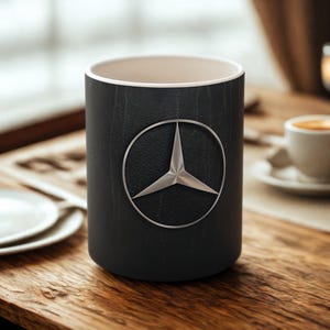 Puede incluir: Taza de café negra con el logotipo plateado de Mercedes-Benz. La taza tiene un aspecto texturizado similar al cuero y un interior crema. Está colocada sobre una mesa de madera.
