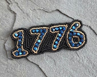 Puede incluir: Un broche bordado con el número "1776". Los números están hechos de cuentas azules, delineadas con cuentas doradas, y sobre un fondo negro de cuentas. El broche se muestra sobre una superficie de piedra gris.
