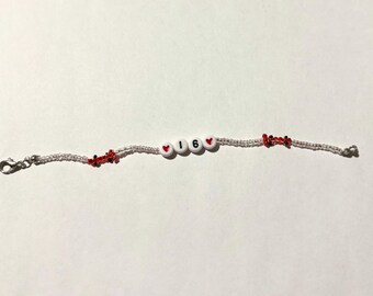 May include: A beaded bracelet with the number "16" spelled out in white letter beads, flanked by red heart beads. The bracelet also features red and black decorative beads and silver clasps.