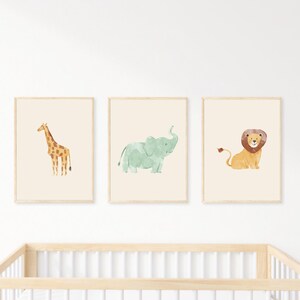 May include: Three framed watercolor prints featuring a giraffe, elephant, and lion. The giraffe is brown and yellow, the elephant is light green, and the lion is tan with a brown mane. The prints are displayed above a wooden crib.
