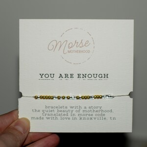 May include: A bracelet with gold and silver beads is displayed on a white card. The card reads "YOU ARE ENOUGH" and includes the text "Morse Motherhood". The card also states the bracelet is translated in morse code.