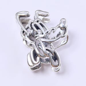 May include: A silver-colored ring with a stylized, abstract design. The ring features a series of interconnected, curved shapes, creating a flowing, sculptural appearance. The metal has a polished finish, reflecting light and adding to the ring's visual appeal.